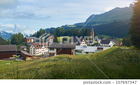 Churwalden village in Switzerland. Formerly Parpan. Beautiful swiss alpine countryside with a medieval bell tower and Rothorn mountain on background. Churwalden village in Switzerland. Formerly Parpan. Beautiful swiss alpine countryside with a medieval bell tower and Rothorn mountain on background. 107697347
