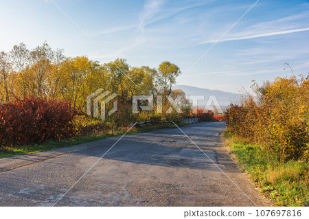 old cracked country road through rural valley in morning light. autumnal scenery with trees in fall colors on the roadside. mountains in the hazy distance old cracked country road through rural valley in morning light. autumnal scenery with trees in fall colors on the roadside. mountains in the hazy distance 107697816