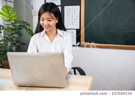 Back to school. Smiling female using computer sitting at school table, Portrait of young woman teacher with laptop at desk in classroom, Online education and learning concept 107698534