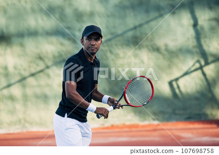 Professional tennis player with racket looking at camera on hard court	 107698758