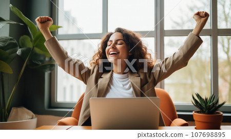 Cute, happy woman with closed eyes sitting at a home office desk with a laptop, raising her arms in a victory pose Cute, happy woman with closed eyes sitting at a home office desk with a laptop, raising her arms in a victory pose 107698895