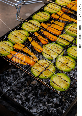 Chopped zucchini and carrots roasting on fire seasoned with aroma herbs and spices. Delicious fresh vegetables grilling on barbecue smoker grid. Diet vegan bbq. Outdoor recreation 107699969