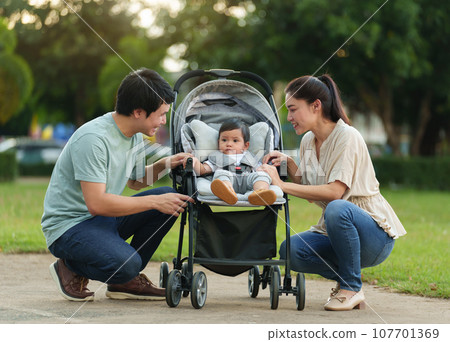 happy parent (father and mother) talking and playing with infant baby in stroller while resting in park happy parent (father and mother) talking and playing with infant baby in stroller while resting in park 107701369