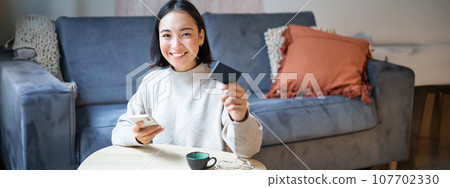 Smiling cute asian woman using credit card and smartphone, paying bills online, holding mobile phone, looking at camera 107702330