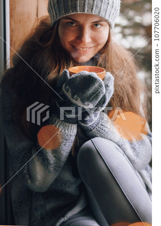 Beautiful  woman holding and drinking a cup of coffee or cocoain in gloves sitting home by the window. Blurred winter snow tree background. Morning, coziness, winter and people concept 107706020
