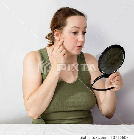 A middle-aged woman looks worriedly at herself in the mirror A middle-aged woman looks worriedly at herself in the mirror 107706081