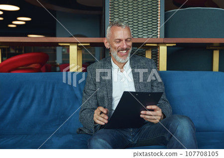 Smiling businessman looking on documents while sitting on sofa in modern office Smiling businessman looking on documents while sitting on sofa in modern office 107708105
