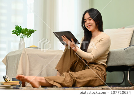 Charming young woman using digital tablet while sitting on floor in living room. Technology and lifestyle concept 107710624