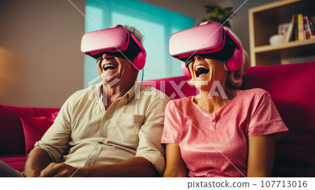Senior mature couple wearing virtual reality headsets sits on the sofa having fun together. Happy retired people using modern VR goggle glasses. New trends and technology concept for all ages. 107713016