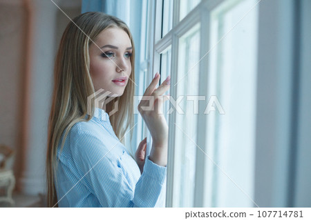 Beautiful woman opening blue curtains looking out window at home Beautiful woman opening blue curtains looking out window at home 107714781