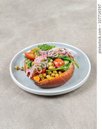 Baked sweet potato with chickpeas, tomatoes, spinach and red onion. Close-up, on a gray background. Baked sweet potato with chickpeas, tomatoes, spinach and red onion. Close-up, on a gray background. 107716597