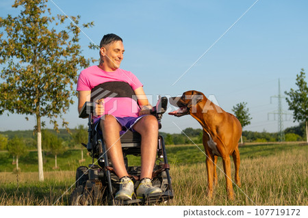 A male in a black wheelchair engages in playtime with his pet dog in a sunlit meadow A male in a black wheelchair engages in playtime with his pet dog in a sunlit meadow 107719172