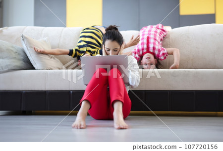 Tired woman with laptop sits on floor with closed palms ears behind on couch children indulge 107720156