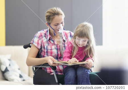 Woman with an oxygen mask and in wheelchair is reading book with little girl 107720201