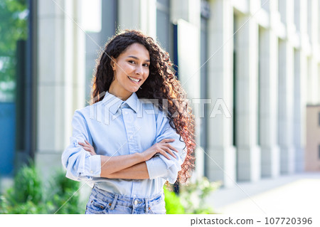 Portrait of happy and successful business woman, boss in shirt smiling and looking at camera inside office with crossed arms, Hispanic woman with curly hair outdoors 107720396