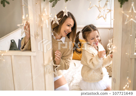 Mother and child laugh merrily playing in the nursery bed with Christmas garland 107721649