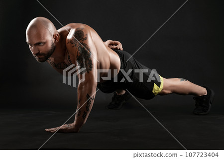 Athletic bald, tattooed man in black shorts and sneakers is posing against a black background. Close-up portrait. Athletic bald, tattooed man in black shorts and sneakers is posing against a black background. Close-up portrait. 107723404