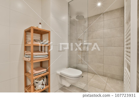 a bathroom with white tile walls and flooring, there is a wooden shelving on the shelf next to the toilet 107723931