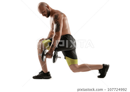 Athletic bald, tattooed man in black shorts and sneakers is posing with a dumbbell isolated on white background. Full length portrait. Athletic bald, tattooed man in black shorts and sneakers is posing with a dumbbell isolated on white background. Full length portrait. 107724992