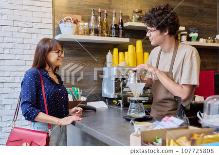 Young male barista talking to woman customer in coffee shop Young male barista talking to woman customer in coffee shop 107725808
