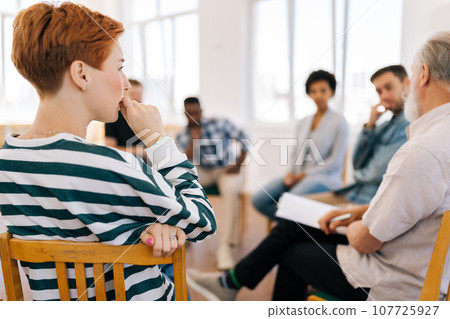 Rear view of thoughtful young woman thinking about mental problem or addiction sitting in circle during group therapy session. Concept of mental health, psychotherapy, depression, social issues. 107725927