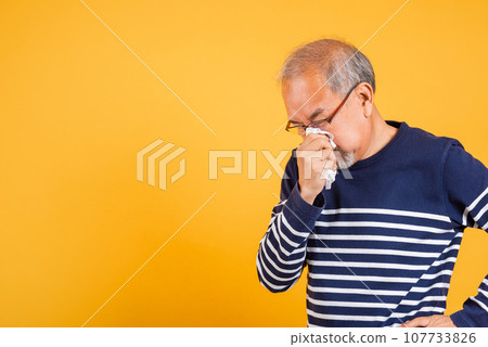 Portrait senior old man blowing nose with tissue studio shot isolated on yellow background, Asian elder man cold having flu and sneezing from sickness virus problem use tissue, pensioner unwell 107733826