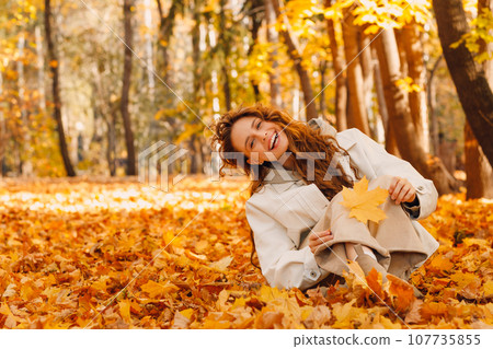 Laughing girl sits in the autumn foliage on the ground of the forest with the yellow leaves Laughing girl sits in the autumn foliage on the ground of the forest with the yellow leaves 107735855