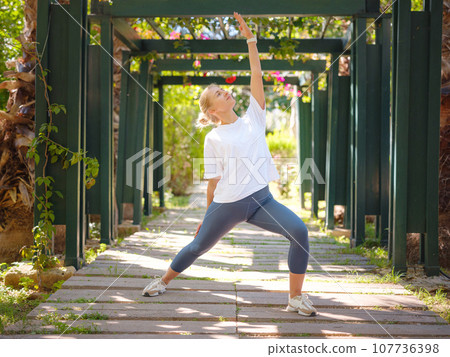 Young woman leading healthy lifestyle and practicing yoga, performs the virabhadrasana exercise, warrior pose, trains alone on summer morning in park. Wellness being concept 107736398