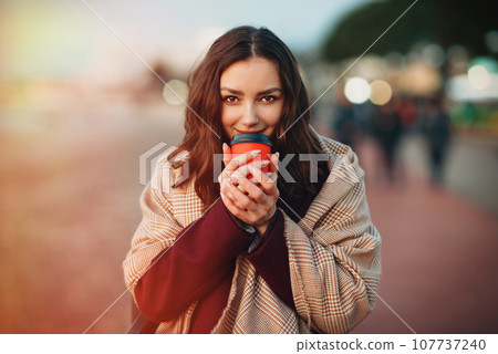 Young smiling woman dressed in autumn coat and plaid drinking hot coffee drink from cardboard cup while strolling outside. Young smiling woman dressed in autumn coat and plaid drinking hot coffee drink from cardboard cup while strolling outside. 107737240