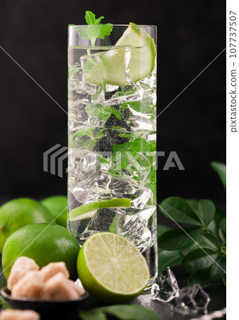 Highball glass of Mojito cocktail with ice cubes,mint and lime on black board with fresh limes Highball glass of Mojito cocktail with ice cubes,mint and lime on black board with fresh limes 107737507