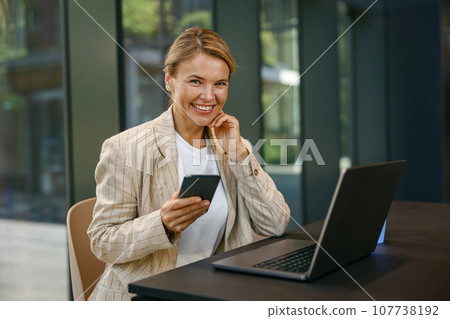 Smiling female entrepreneur use phone while working on laptop in modern office background  107738192