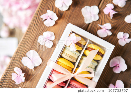 Dessert French Macarons in present box. Sweet dessert on a flowers background. Still life with macaroni cakes and different flower. Delivery concept, coffee and flower shop Dessert French Macarons in present box. Sweet dessert on a flowers background. Still life with macaroni cakes and different flower. Delivery concept, coffee and flower shop 107738797