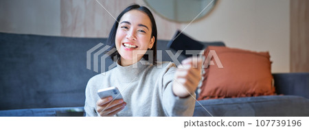Smiling asian girl with smartphone and credit card, does her shopping online, uses mobile phone to order home delivery Smiling asian girl with smartphone and credit card, does her shopping online, uses mobile phone to order home delivery 107739196