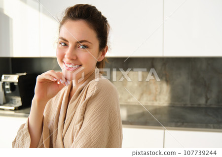 Portrait of beautiful and happy woman standing in kitchen in bathrobe, posing and smiling at camera, cooking food, preparing meal, eating lunch near counter 107739244