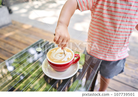 Cup of hot cappuccino. Little girl stir sugar into cappuccino. Breakfast at the bakery cafe. A cup of coffee on the veranda on a sunny day. Coffee break at the cafe. Close-up 107739501