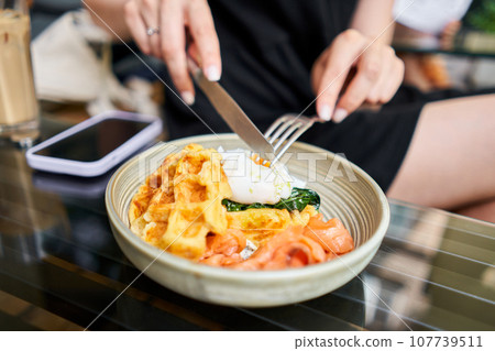 Woman cuts poached egg and yolk flows out. Potato waffles, avocado cream with salmon and egg . Healthy breakfast, protein. Restaurant dish.  107739511
