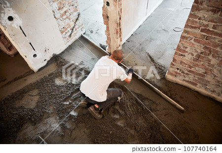 Male construction worker placing screed rail on the floor covered with sand-cement mix. Man smoothing and leveling surface with straight edge while screeding floor in apartment. 107740164