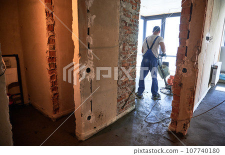 Worker using troweling machine while screeding floor in apartment under renovation. Man finishing concrete surface with floor screed grinder machine in room with large window. 107740180