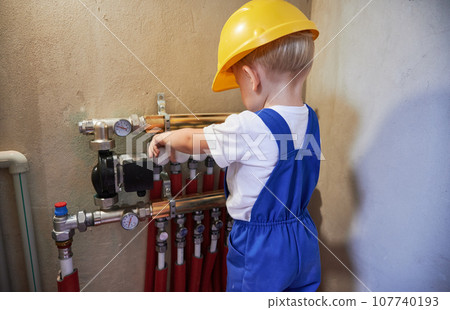 Baby boy checking plumbing installation with pipes and thermometer gauge. Kid plumber in workwear working with plumbing pipe system in apartment under renovation. Baby boy checking plumbing installation with pipes and thermometer gauge. Kid plumber in workwear working with plumbing pipe system in apartment under renovation. 107740193
