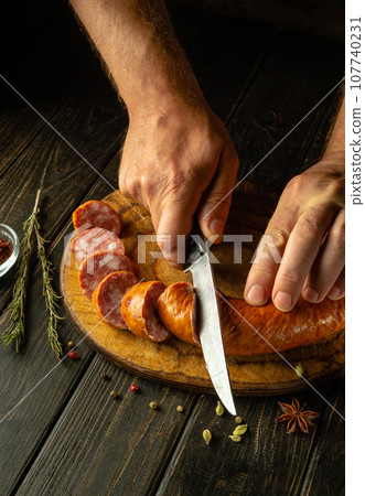 The chef slice a meat veal sausage with a knife on a wooden cutting board. The concept of preparing delicious sandwiches for breakfast on the kitchen table 107740231