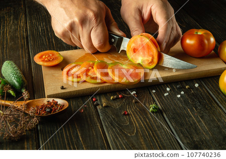 Sliced tomatoes for a vegetable dish. Knife in the chef hand while slicing a tomato on a kitchen board Sliced tomatoes for a vegetable dish. Knife in the chef hand while slicing a tomato on a kitchen board 107740236