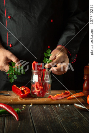 Sliced sweet red peppers on a wooden cutting board, a professional cook in a black uniform prepares a salad. A man's hand puts parsley into a jar 107740252