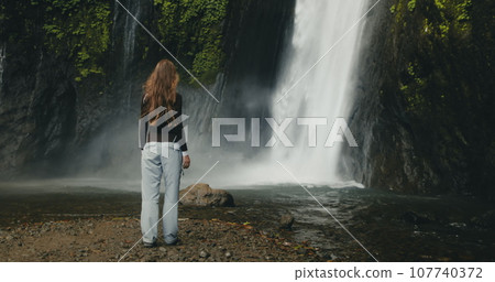 Tourist girl raising arms standing at powerful waterfall Air Terjun Munduk, Indonesia, Bali island. Woman posing, back view. Beautiful nature landscape. Travel, tourism, holiday 107740372