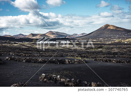 Traditional white houses in black volcanic landscape of La Geria wine growing region with view of Timanfaya National Park in Lanzarote. Touristic attraction in Lanzarote island, Canary Islands, Spain Traditional white houses in black volcanic landscape of La Geria wine growing region with view of Timanfaya National Park in Lanzarote. Touristic attraction in Lanzarote island, Canary Islands, Spain 107740765