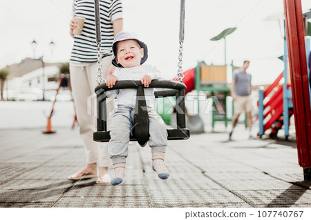 Mother pushing her infant baby boy child on a swing on playground outdoors Mother pushing her infant baby boy child on a swing on playground outdoors 107740767