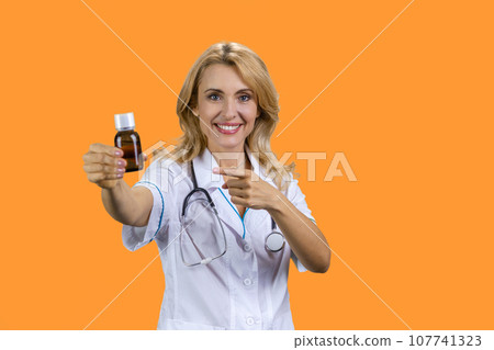 Portrait of happy blonde mature female doctor pointing at medicine bottle. Isolated on orange. Portrait of happy blonde mature female doctor pointing at medicine bottle. Isolated on orange. 107741323