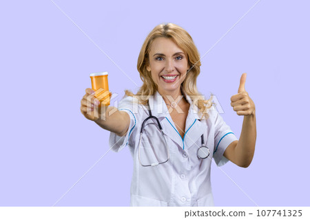 Portrait of happy caucasian female doctor holding a can of pills and gives her thumb up. Isolated on purple. Portrait of happy caucasian female doctor holding a can of pills and gives her thumb up. Isolated on purple. 107741325