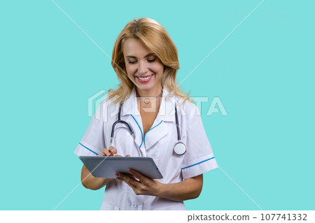 Portrait of cheerful blonde mature medical doctor woman using tablet device. Isolated on turquoise. Portrait of cheerful blonde mature medical doctor woman using tablet device. Isolated on turquoise. 107741332