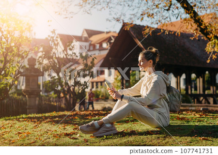 Young Fashionable Teenage Girl With Smartphone In Europian Park In Autumn Sitting At Smiling. Trendy Young Woman In Fall In Park Texting. Retouched, Vibrant Colors. Beautiful Blonde Teenage Girl Young Fashionable Teenage Girl With Smartphone In Europian Park In Autumn Sitting At Smiling. Trendy Young Woman In Fall In Park Texting. Retouched, Vibrant Colors. Beautiful Blonde Teenage Girl 107741731