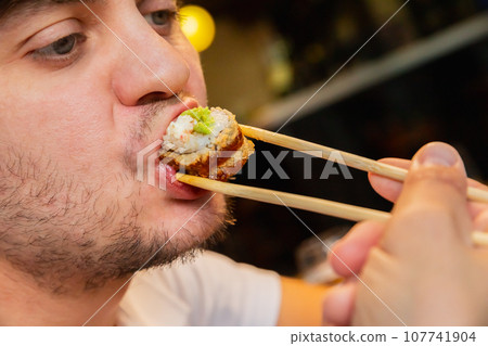 Young adult male man eating baked Japanese sushi roll with bamboo chopsticks at sushi restaurant. Tempura fried roll and wasabi sauce. Male unshaven face with roll in his mouth close-up. Seafood. 107741904